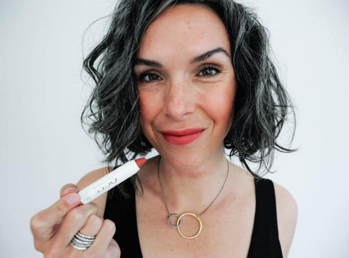This Organic Girl applying OGEE Sculptured Lip Oil while looking into the camera wearing a black tank dress against a white background