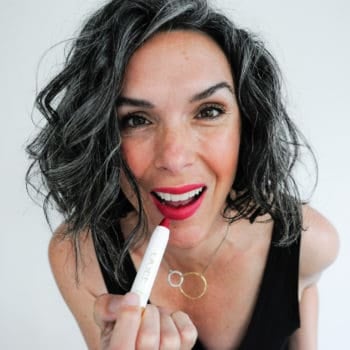 This Organic Girl applying OGEE Sculptured Lip Oil while looking into the camera wearing a black tank dress against a white background