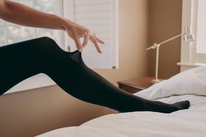 A woman's leg is poised up on the side of her bed in black tights.