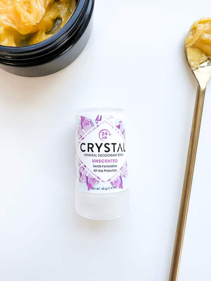 A stick of crystal deodorant lays next to a scoop full of yellow body balm in a gold spoon.