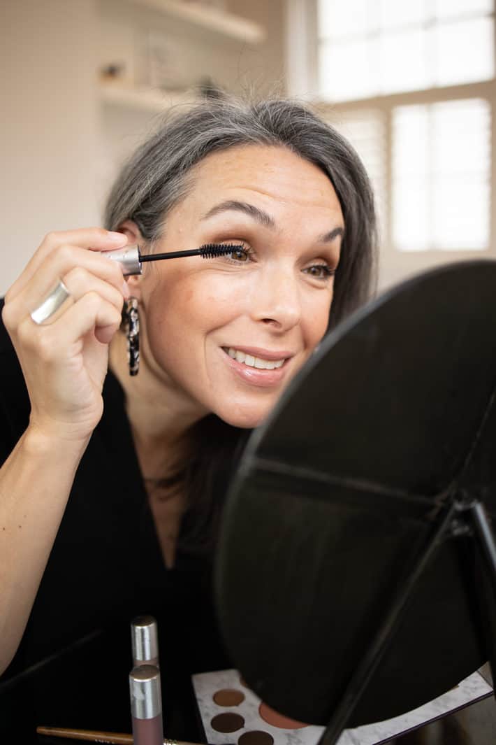 a woman smiling as she applies mascara to her eyelashes