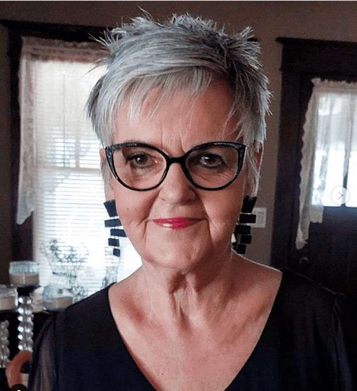 a woman with spiky gray hair and black glasses looks into the camera