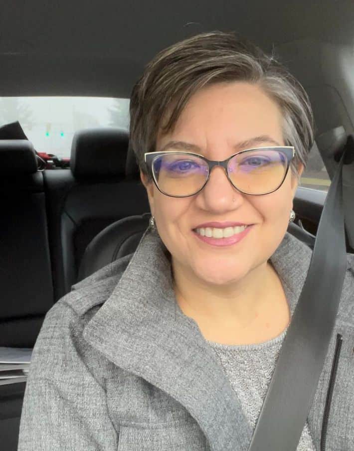 a woman in glasses with pink lipstick sits in her car and smiles at the camera