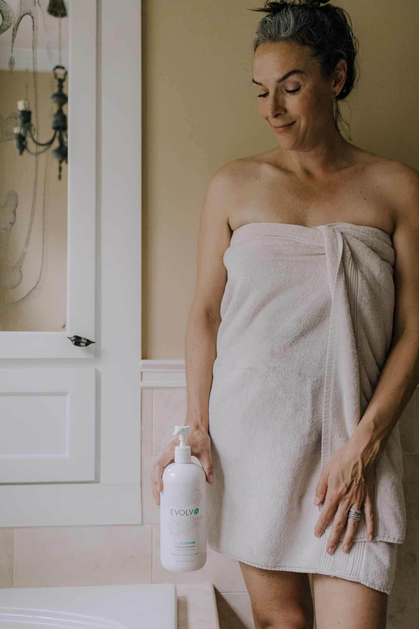 a woman holds a bottle of evolvh shampoo near her bathtub