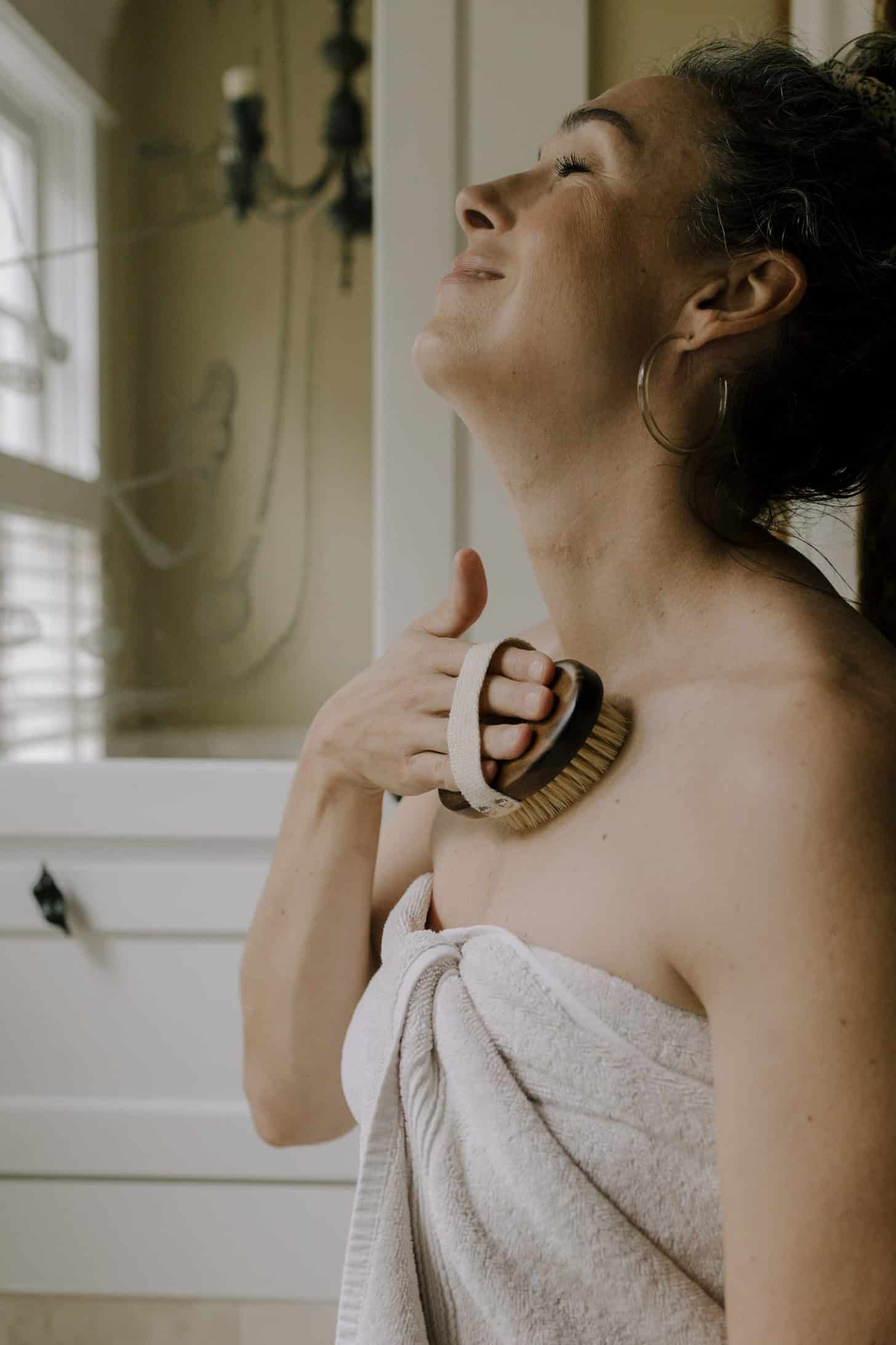 a woman dry brushes her skin while wearing a towel