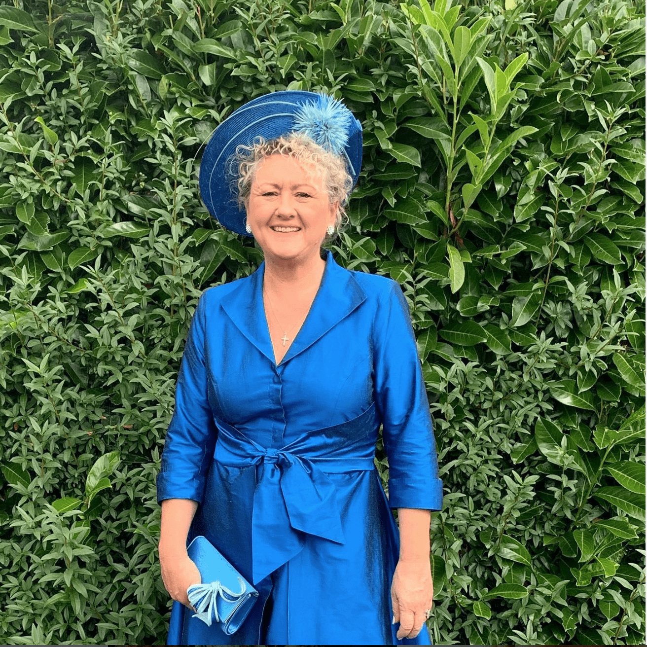 a woman in a blue dress and hat stands in front of a wall of greenery