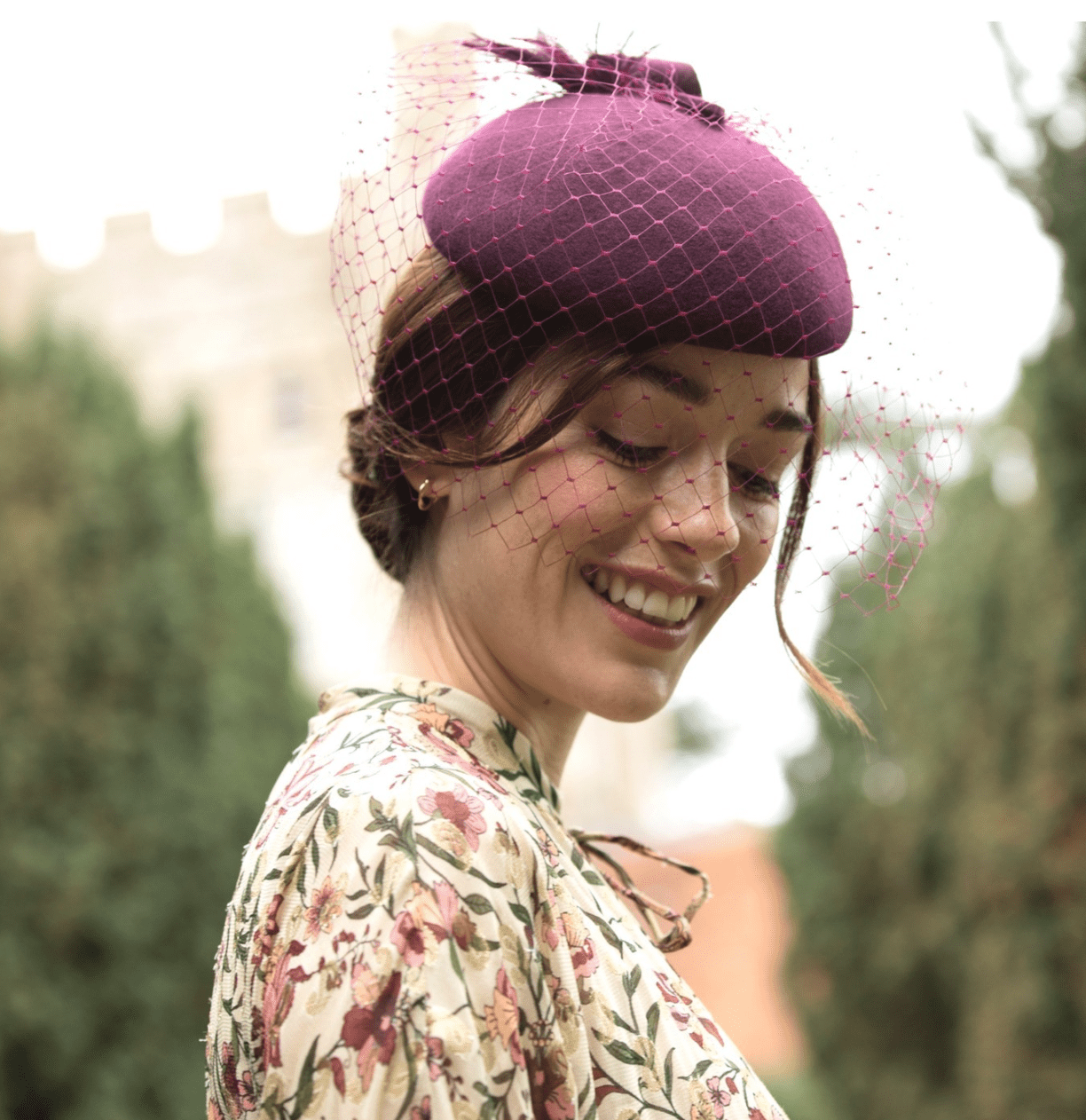 a woman with a raspberry pillbox hat looks down towards her floral dress