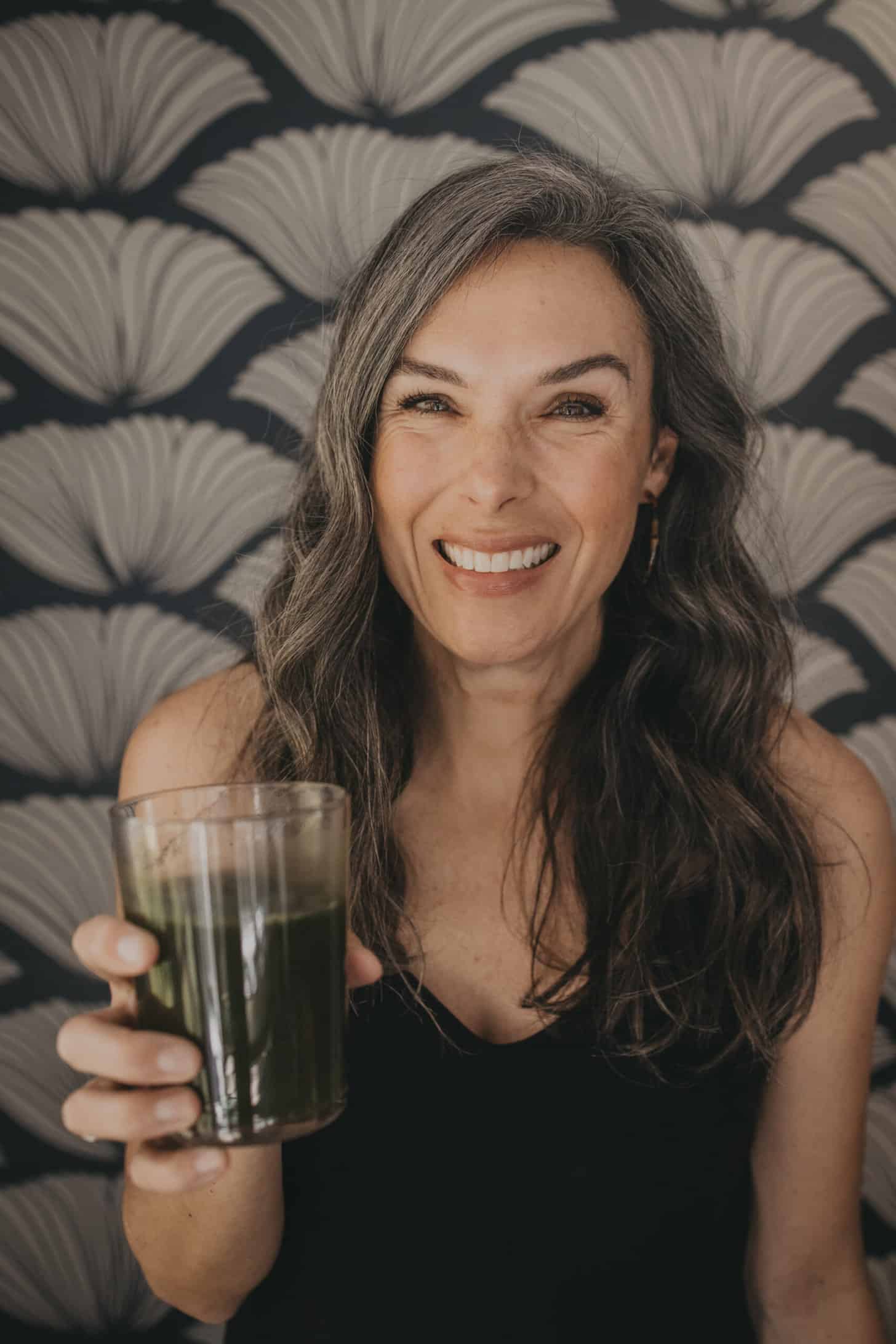 a woman with long wavy hair holds up a glass of green juice