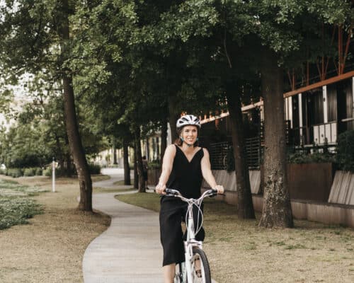 a woman in a black jumpsuit rides a bike down a tree lined path