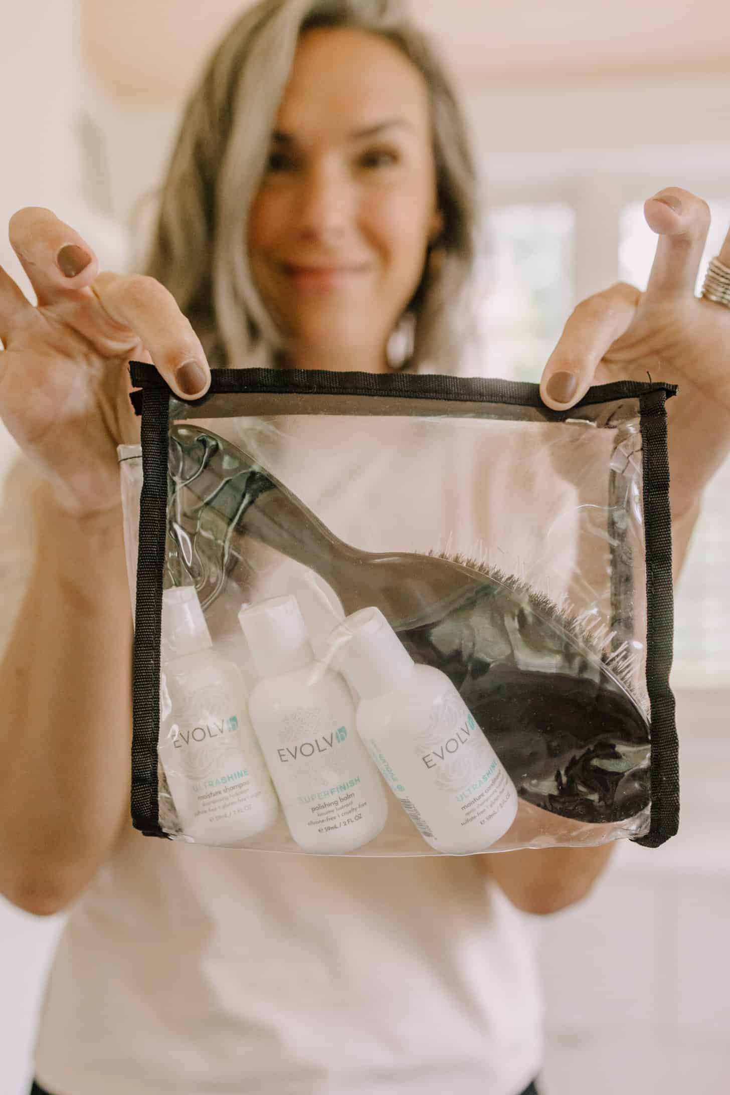 a woman holds up a small bag of evolvh travel sized hair care products and a brush