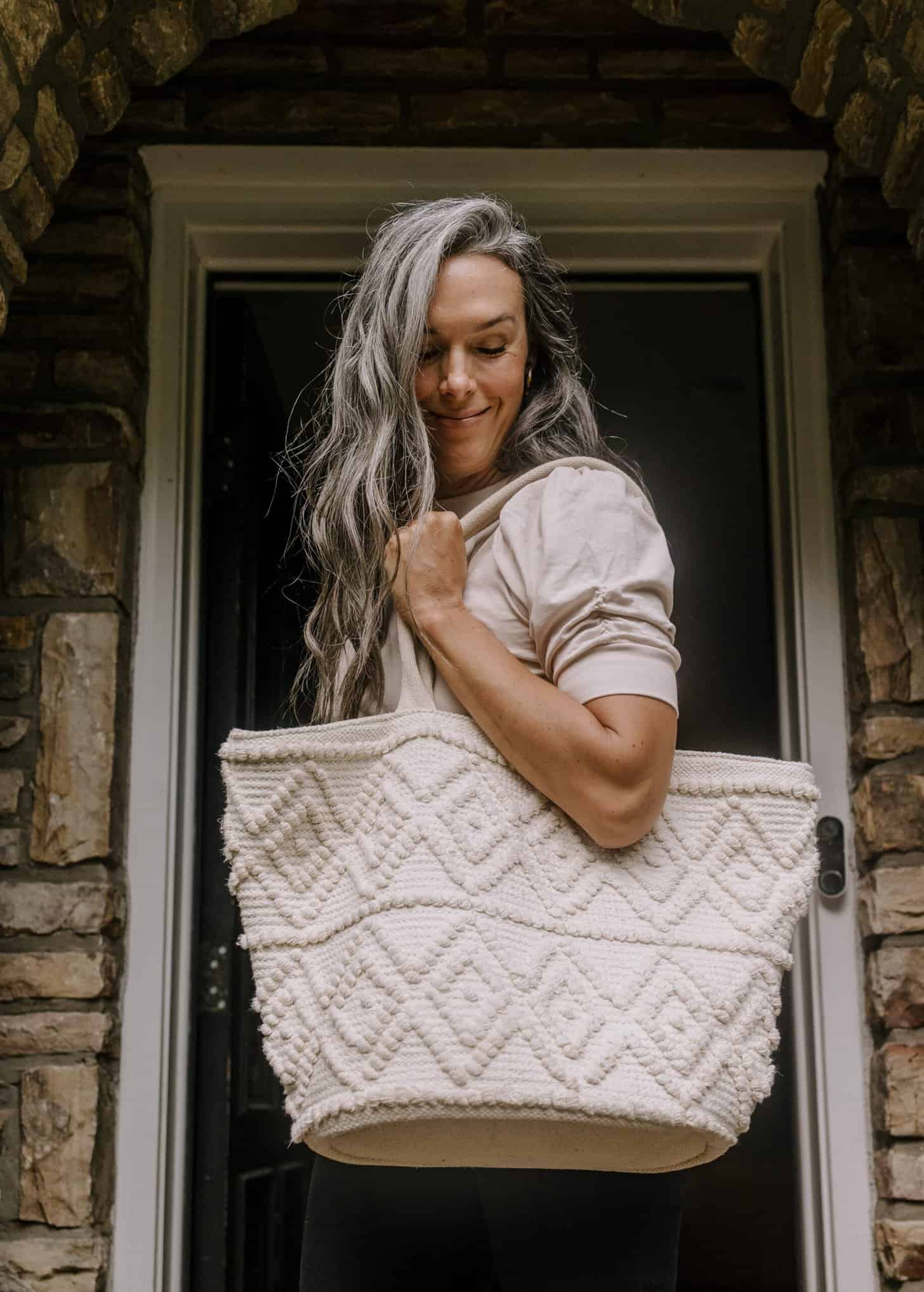 a woman stands outside a door carrying a large woven bag