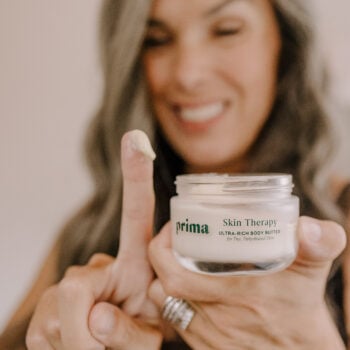 a woman holds up a tub of Prima Skin Therapy along with glob of it on her finger