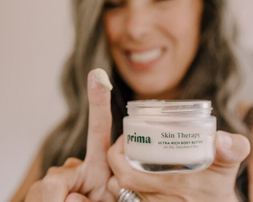 a woman holds up a tub of Prima Skin Therapy along with glob of it on her finger